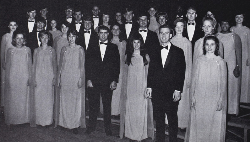 A group of about 30 students standing on a stage wearing formal gowns and suits.