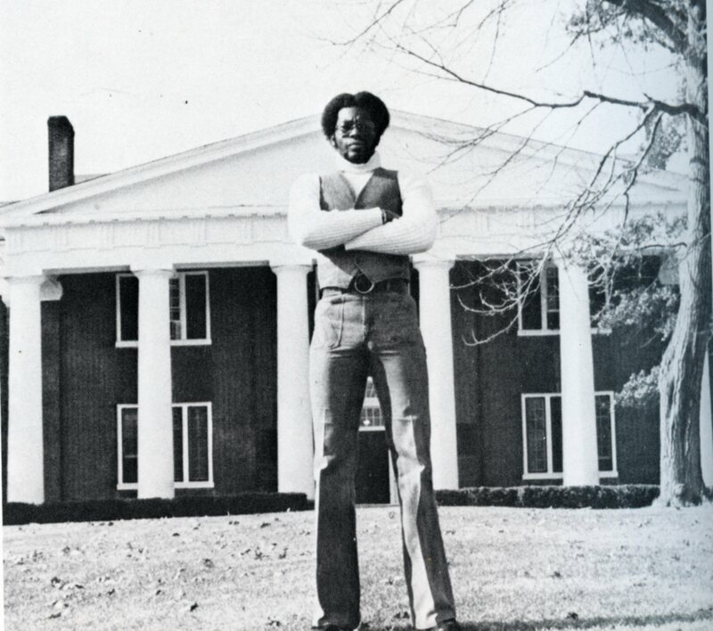 Titus Bryant stands with arms folded across chest on the lawn in front of a two story brick building with white columns. The angle of the image makes him appear larger than the building.
