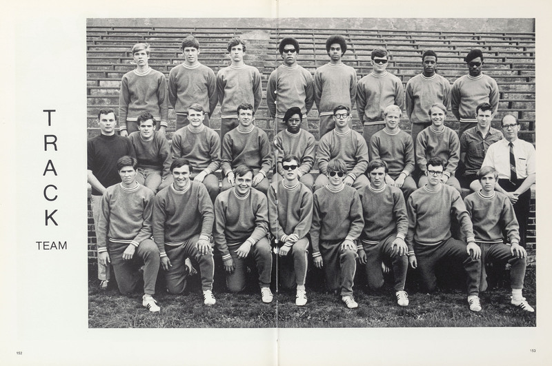 Official track team photograph. Members wear matching track suits. They are arranged in three rows in front of bleachers. Coach stands to  right.