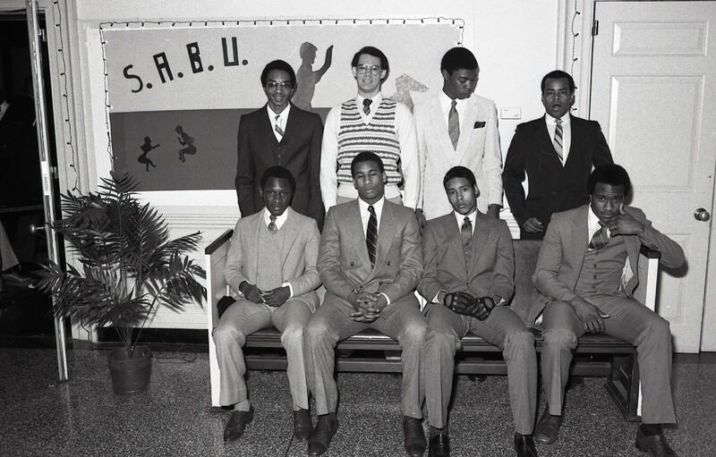 Eight male students in front of a SABU poster, four seated on a couch, four standing behind them. All men are in suits.