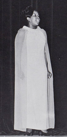 Sarah Reese, standing on stage in a white gown, singing