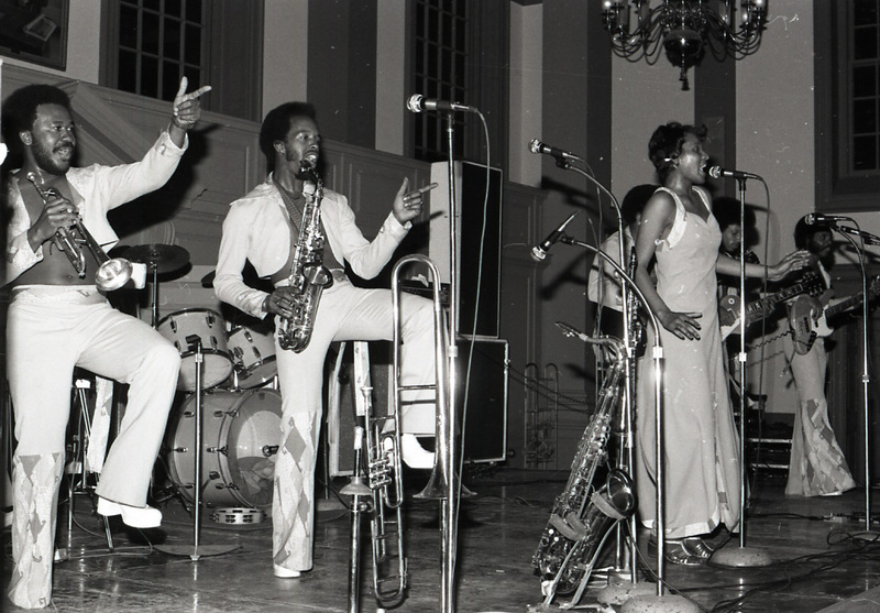 Six performers on a stage, a saxaphone player, trumpet player, singer, two guitarists, and a backup singer. The drummer is not in the image but the drum kit is. The trumpet player and saxophone player are dancing and pointing at the crowd while the lead singer is singing into the microphone.