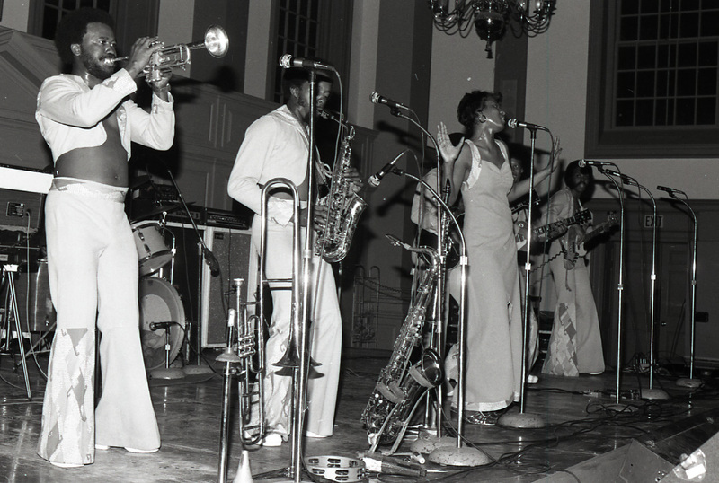 Six performers on a stage, a saxaphone player, trumpet player, singer, two guitarists, and a backup singer. The drummer is not in the image but the drum kit is. The trumpet player and saxophone player have their instruments to their mouths.