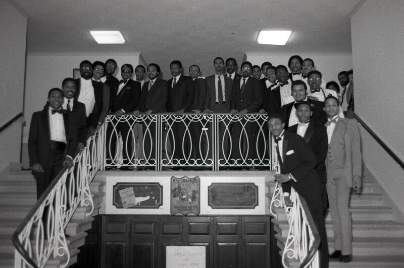 A large group of Black men in formal attire posing on staircase.